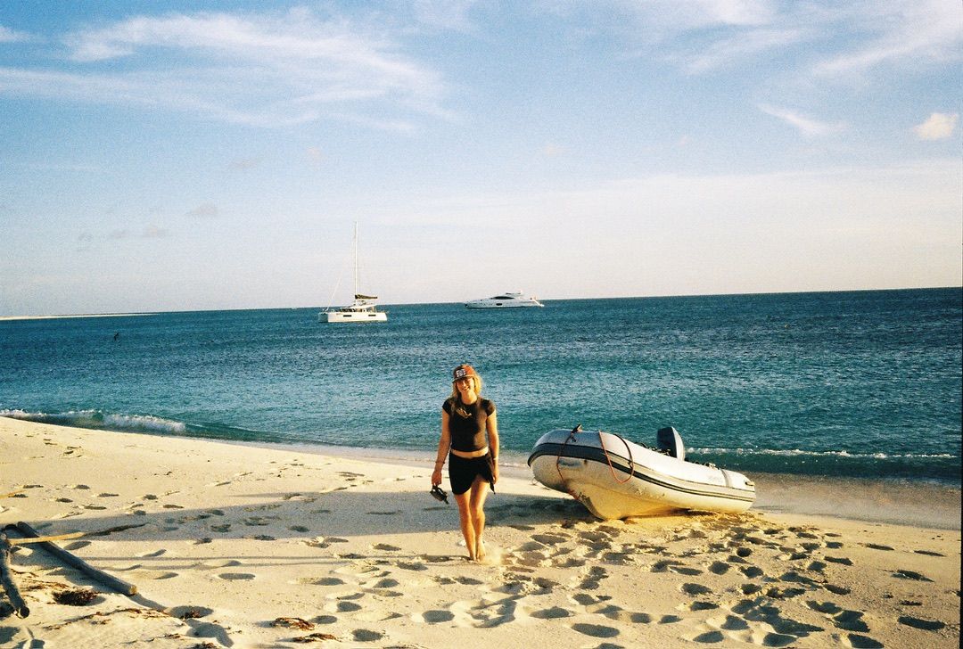 Crew member walking on a white sand beach with Laurelli and the dinghy anchored offshore