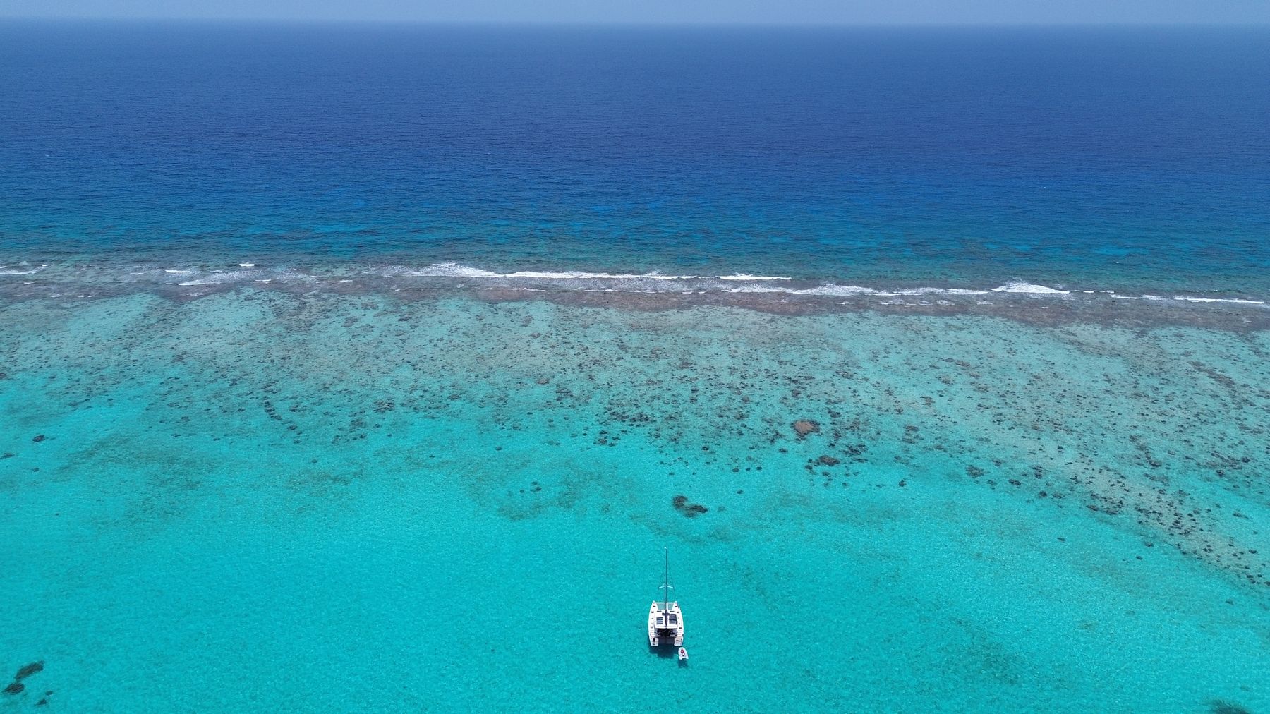Aerial view of Laurelli anchored alone at the edge of a reef, solar panels visible on the roof